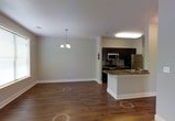 This single-floor residence offers a clean, open-concept design that integrates the living, dining, and kitchen areas into one cohesive and functional space. Rich, dark hardwood flooring runs throughout, providing a warm contrast to the neutral gray walls and bright white trim and baseboards. The dining area is anchored by an elegant pendant chandelier with multiple lights, adding a refined touch to the otherwise understated interior. Natural light filters in through a well-placed window, further brightening the open layout. The kitchen is outfitted with dark espresso-toned cabinetry that extends to the ceiling, paired with stainless steel appliances including a microwave and range. A peninsula with a dark stone countertop provides additional prep space and serves as a natural divider between the kitchen and the living areas. The overall ambiance of the space is modern and practical, with a neutral color palette that lends itself well to a variety of furnishing styles. The seamless flow between areas makes the space feel open and inviting, while thoughtful design details like recessed lighting and visible ventilation contribute to its functional appeal.