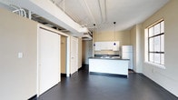 This single-floor space showcases an open-concept design with distinct industrial character. The layout integrates a spacious main living area with dark-stained flooring that extends throughout, creating a cohesive and grounded aesthetic. Exposed ceiling infrastructure, including visible ductwork, piping, and structural elements, contributes to the space's loft-like industrial ambiance.

The kitchen is positioned as a central feature, incorporating a white peninsula with a dark countertop, a double-basin sink, and white upper cabinetry. A full-size refrigerator anchors one end of the kitchen, while two blue pendant lights hang overhead, adding a distinct visual accent against the neutral tones of the walls and cabinetry.

Adjacent to the main living area, a cluster of doors suggests access to private rooms, including a bedroom and bathroom. The bathroom, visible in the floor plan, includes standard fixtures. Louvered closet doors and standard-height interior doors define the private spaces without interrupting the open flow of the main area.

The high ceilings amplify the sense of volume throughout, while the warm beige wall tones balance the rawness of the exposed overhead elements, resulting in a functional yet characterful space.