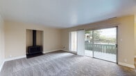 This single-floor residence offers a thoughtfully laid-out living space with a warm, neutral color palette throughout. The generous living room serves as the heart of the home, featuring plush carpeting, a freestanding wood-burning stove, and large sliding glass doors that open onto a private balcony with views of surrounding greenery — filling the room with natural light and a connection to the outdoors. An additional carpeted room with its own sliding door to the balcony provides flexible use as a bedroom or sitting area, complete with a built-in sliding-door closet for ample storage. The galley-style kitchen is efficiently designed with white cabinetry, dark granite-style countertops, light wood-look flooring, and white appliances including a range and refrigerator. A hallway with built-in shelving connects the rooms, leading to a well-appointed bathroom featuring a soaking tub with shower, dark stone countertop vanity with white cabinetry, a large mirror, and wood-look flooring. A second vanity area is accessible from the hallway, offering added convenience. The consistent use of warm beige tones, clean white trim, and wood-look flooring in the wet areas creates a cohesive and inviting atmosphere throughout the space.