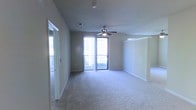 This single-floor space centers around a spacious, open living area finished with wall-to-wall carpeting in a neutral tone, contributing to a clean and understated aesthetic. The room is equipped with a ceiling fan, providing both functional air circulation and a subtle design element overhead. Smooth, light-toned walls are complemented by white baseboards, creating a cohesive and simple interior palette.

Natural light is a defining feature of the space, streaming in through a large floor-to-ceiling window and sliding glass door that opens onto a private balcony. The balcony, framed by a metal railing, offers an outdoor extension of the living area and adds depth to the room's overall feel.

To one side, a mirrored wall panel enhances the sense of openness by reflecting light throughout the room. On the opposite side, a doorway with a white trim frame leads to an additional area, likely a closet or adjoining room, also featuring a mirrored sliding door. Ceiling-mounted smoke detector and a standard HVAC vent are discreetly integrated into the ceiling. Overall, the space presents a bright, functional, and move-in-ready environment.