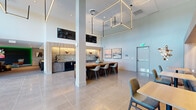 Located at 4140 Jason St in Denver's vibrant LoHi neighborhood, this contemporary amenity space delivers a well-appointed, multi-functional environment across a single floor. The main gathering area impresses with soaring double-height ceilings, large-format polished tile flooring, and an eye-catching geometric cube light fixture that anchors the open layout. A standout communal kitchen and lounge area features a dramatic live-edge island countertop with turquoise resin accents, surrounded by modern upholstered seating. The kitchen wall is lined with tastefully illuminated open shelving, textured tile backsplash, and dark cabinetry housing a beverage cooler and microwave. Curated sculptural wall art adds a gallery-like character throughout. A built-in banquette nook with custom lighting provides an intimate co-working or dining retreat. Expansive glass entry doors flood the space with natural light and offer views of an adjacent outdoor area. The entertainment lounge, accessible through the main space, features ambient green LED accent lighting, a large wall-mounted screen, comfortable sectional seating, and a shuffleboard table — creating a dynamic social setting. The overall ambiance blends modern sophistication with approachable comfort, ideal for community gatherings and social events.