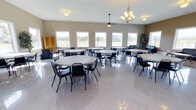 This banquet center offers a spacious, open-plan event hall designed to accommodate a variety of gatherings. The room is furnished with multiple round tables and black stackable chairs arranged throughout, providing flexible seating configurations for guests. The polished terrazzo flooring reflects light beautifully, enhancing the bright and welcoming atmosphere of the space.

Natural light floods the hall through a row of large windows along one wall, offering views of the surrounding outdoor landscape. Overhead, a decorative chandelier serves as a focal point, complementing a ceiling fan that adds both style and airflow to the room. The neutral-toned walls contribute to a clean, versatile backdrop suitable for a wide range of events.

Along the perimeter, additional seating areas with sofas and armchairs provide informal gathering spots, while a wooden armoire adds a touch of warmth to the décor. A potted tree near the windows introduces a natural element to the interior. The single-floor layout ensures easy accessibility throughout the venue, making it a practical and attractive choice for banquets, receptions, and community events.