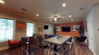 This sports lounge is a well-appointed, single-floor gathering space designed for both relaxed lounging and lively social interaction. The interior is defined by a bold color palette, with striking burnt-orange accent walls contrasting against cool gray tones, creating an energetic yet comfortable atmosphere. Crown molding and recessed lighting run throughout, lending the space a polished, finished look, while dark hardwood-style flooring anchors the design.

The lounge is organized into distinct zones. A comfortable seating area features a large navy sectional sofa, a modern coffee table atop a patterned area rug, and a sculpted lounge chair with ottoman — all oriented toward a large wall-mounted flat-screen TV. Framed abstract artwork decorates the walls throughout, adding a curated, contemporary feel.

A separate dining and game area offers high-top tables with sleek bar-height chairs, a foosball table, and a mini refrigerator — perfect for casual entertaining. An additional wall-mounted TV and built-in booth-style seating with blue upholstery round out this zone.

Ceiling fans provide comfort alongside the recessed lighting, and natural light filters in through slatted blinds on the windows. Decorative potted plants are placed throughout the space, adding a touch of greenery to the vibrant interior.