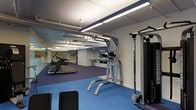 This fitness center offers a comprehensive workout environment designed to accommodate a range of exercise needs. The space is finished with durable, color-zoned flooring — blue in the central training area and purple beneath the cardio equipment — providing both visual organization and functional cushioning underfoot. A row of treadmills lines one side of the room, facing full-length mirrors that visually expand the space and allow users to monitor their form during exercise. Overhead, exposed ductwork and bright linear fluorescent lighting run the length of the ceiling, giving the space an industrial yet clean aesthetic. The strength training area features a multi-function cable machine with weight stacks and a freestanding captain's chair for core and upper-body exercises. The mirrored walls throughout further enhance the sense of openness while keeping the environment focused and functional. The layout is thoughtfully arranged to allow ample circulation between equipment, ensuring a safe and comfortable workout experience. Overall, the space presents a practical and well-maintained fitness facility suited for both cardiovascular and resistance training.