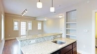 This single-floor space showcases a thoughtfully designed open-concept layout that integrates the kitchen and living area into one cohesive environment. The kitchen is anchored by an expansive granite-topped island with light, speckled patterning and a stainless steel undermount sink. Dark cabinetry beneath the island provides a striking contrast to the stone surface. Two cylindrical pendant lights hang above the island, offering focused task lighting while contributing to the overall aesthetic. A set of built-in open shelving units to the right of the kitchen offers convenient display and storage space. The living area beyond features neutral taupe walls, flush ceiling-mounted lighting, and a track lighting fixture for adjustable illumination. Multiple windows, including a set of glass-paneled double doors, allow natural light to fill the space and create an airy, open atmosphere. The seamless flow between the kitchen and living zone makes this layout well-suited for both everyday living and entertaining, while the clean finishes and layered lighting contribute to a polished, functional ambiance throughout.