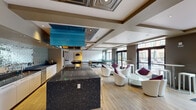 This thoughtfully designed single-floor amenity space blends modern sophistication with eclectic flair across several distinct zones. The centerpiece is a gourmet kitchen anchored by a generous dark granite island with bar seating, complemented by sleek white cabinetry and a striking mirrored mosaic tile backsplash that catches and reflects light throughout the space. A bold turquoise range hood suspended from an ornate coffered ceiling panel serves as a dramatic focal point, adding an artistic contrast to the otherwise refined kitchen design.

Adjacent to the kitchen, an open lounge area features contemporary white swivel chairs with plum accent cushions, glass-topped tables, and light hardwood flooring, all bathed in natural light streaming through floor-to-ceiling windows and glass doors that connect the interior to an outdoor area. A billiards table occupies its own dedicated zone, ideal for recreation and social gatherings.

At the far end of the space, a more intimate lounge area exudes warmth and character, highlighted by a grand white ornate fireplace, bold magenta and gold textured wall treatments, and a richly upholstered sofa layered with colorful throw pillows. A wall-mounted flat-screen TV and stylish media console complete this cozy retreat. Throughout, coffered and recessed ceilings add architectural depth and dimension to the entire space.