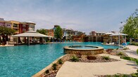 This expansive resort-style outdoor amenity area is designed to deliver a full leisure experience centered around a generously sized, freeform lagoon pool with vivid turquoise water. A sandy beach shoreline lined with colorful lounge chairs and palm trees gives the space a distinctly tropical feel, evoking a beachfront retreat. A dramatic stone waterfall feature anchors one end of the pool, while a raised circular spa sits adjacent, crafted from natural stone and surrounded by decorative rock landscaping. The pool deck extends around the entire perimeter, offering ample space for sunbathing under large market umbrellas. A covered outdoor pavilion provides a shaded gathering area equipped with ceiling fans, wall-mounted televisions, and modern white communal tables with contemporary seating — ideal for group activities or casual relaxation. Nearby, a decorative stone-paved courtyard features a long ornamental fountain as its centerpiece, framed by manicured landscaping including palms, ornamental grasses, and seasonal plantings. Teal-accented architectural columns and crisp white trim throughout the pavilion and surrounding structures tie the design together with a coastal aesthetic. The entire complex enjoys open sky views and abundant natural light, creating a vibrant, resort-caliber outdoor living environment.