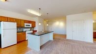 Located at 610 Towne Center Dr in Joppa, Maryland, this single-floor, one-bedroom, one-bathroom residence offers a thoughtfully designed layout that maximizes comfort and functionality. The open-concept kitchen and living area forms the heart of the home, where warm hardwood-style flooring in the kitchen transitions seamlessly into plush carpeting in the living space. The kitchen is outfitted with honey-toned wood cabinetry, a tile backsplash, white appliances, and a central island with a dark countertop and pendant lighting overhead — ideal for both cooking and casual dining. Large windows allow generous natural light to fill the living area, creating a bright and welcoming atmosphere. The bedroom is a comfortable retreat, featuring plush carpeting, ample wall space, and direct access to a generously sized walk-in closet equipped with wire shelving on multiple levels. The full bathroom includes a tiled tub-and-shower combination, a vanity mirror, and durable tile flooring. A dedicated laundry area houses a stacked washer and dryer unit alongside a water heater, adding everyday convenience. The overall design is clean, functional, and well-suited for comfortable single-level living.