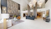 Located at 320 Providence Main St NW in Huntsville, Alabama, this entrance lobby is a striking example of contemporary design executed with premium materials and careful attention to architectural detail. The single-floor space encompasses a generously proportioned footprint that immediately conveys a sense of grandeur upon entry.

The lobby's most commanding feature is its soaring double-height ceiling, which amplifies the sense of volume throughout the space. Anchoring one wall is a dramatic floor-to-ceiling slab of richly veined marble in deep grays and browns, paired with a built-in fireplace and flanking shelving niches. Opposite this focal point, cascading crystal-and-glass chandeliers descend from the ceiling, providing luminous visual interest at multiple levels.

The reception desk, finished in dark charcoal tones, is framed by a tall decorative bronze lattice screen, creating a refined and functional centerpiece. Large black-framed windows and glass entry doors flood the space with natural light while offering views of the surrounding streetscape. Light-toned large-format tile flooring unifies the entire area, complemented by a curated seating arrangement featuring neutral upholstery and warm accent furnishings.

Adjacent to the main lobby, a dedicated mailroom with herringbone-patterned tile flooring houses extensive built-in postal boxes, finished with warm wood cabinetry and accented by a brass pendant fixture.