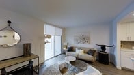 This single-floor space is centered around a thoughtfully designed living area that balances modern aesthetics with a warm, inviting atmosphere. The room features a clean, neutral color palette of whites and soft beiges, complemented by light hardwood-style flooring that runs throughout the space.

The living area is anchored by a comfortable beige sofa adorned with an assortment of decorative pillows in muted tones. A textured, shaggy area rug defines the seating zone, where a round white side table and an organic-shaped coffee table serve as functional yet stylish centerpieces. A wicker accent chair adds a natural, earthy element to the composition.

Floor-to-ceiling sliding glass doors flood the room with natural light and provide direct access to an outdoor area, enhancing the sense of openness. A roller shade offers privacy and light control as needed. The walls are dressed minimally, with a notable large-format print depicting a Parisian cityscape, adding an artistic focal point to the room.

Lighting is carefully considered, featuring a sculptural arc floor lamp alongside a classic table lamp, creating layered illumination. Additional decorative accents, including a dark sculptural figurine, contribute to the space's curated, contemporary character.