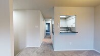 This single-floor residence offers a clean, functional layout designed for comfortable everyday living. The open living and dining area is generously sized, finished with soft wall-to-wall carpeting and neutral tones throughout, and anchored by an elegant chandelier that adds a warm, inviting ambiance. A pass-through opening connects the living space to the kitchen, maintaining an open feel while defining each area.

The kitchen stands out as a highlight of the unit, featuring crisp white shaker-style cabinetry with gold-toned hardware, dark granite countertops, a hexagonal tile backsplash, and stainless steel appliances including a dishwasher and electric range. A matte black pull-down faucet over the undermount sink adds a modern, contrasting accent. A skylight overhead bathes the kitchen in natural light.

The bedrooms are spacious and carpeted, each offering a calm, neutral atmosphere. Windows allow natural light to filter in, and one bedroom includes a louvered closet door for storage. A wall-mounted HVAC unit and baseboard heating ensure year-round comfort. The bathroom is simply finished with wood-look flooring, a white vanity, and a full tub-shower combination. A hallway connects all rooms, providing a cohesive and easy-to-navigate floor plan.