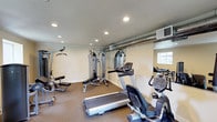 This dedicated fitness space offers a comprehensive workout environment designed to accommodate a range of exercise routines. The room is outfitted with a solid selection of equipment, including a treadmill, stationary bike, and multiple cable and weight machines, making it suitable for both cardiovascular and strength training workouts. The layout is thoughtfully arranged to maximize usable floor space, with carpeted flooring providing comfort underfoot throughout the room. A full-length mirror along one wall enhances the sense of openness while allowing users to monitor their form during exercise. Recessed ceiling lights provide bright, even illumination across the entire space, complemented by natural light filtering in through a window along one wall. Exposed ductwork along the upper portion of the wall adds an industrial touch to the otherwise clean and neutral aesthetic, with warm beige walls contributing to an inviting atmosphere. A wall-mounted HVAC unit ensures climate control for year-round comfort. The single-floor layout keeps everything easily accessible, making this a practical and well-appointed facility for daily fitness use.