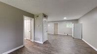 This single-floor residence offers a practical and well-organized layout designed for comfortable everyday living. The open living area serves as the heart of the home, featuring durable wood-look vinyl plank flooring in a warm gray tone and a large window that frames outdoor greenery, filling the space with natural light. Adjacent to the living area, the kitchen is equipped with ample white cabinetry, dark laminate countertops in an L-shaped configuration, a gas range, a refrigerator, and a double sink — providing generous storage and workspace. Multiple bedrooms are finished with neutral-toned carpet and offer access to natural light through well-placed windows; one bedroom includes a built-in closet with a shelving rod for added storage convenience. The full bathroom is clean and functional, featuring wood-look tile flooring, a white vanity with cabinet storage, and a tub-shower combination. Throughout the home, walls are painted in a consistent warm gray palette, complemented by crisp white trim and panel-style doors. A louvered closet provides additional storage in the common area, and HVAC vents are integrated throughout for year-round climate control. The textured ceilings and thoughtful layout contribute to a tidy, move-in ready atmosphere.