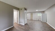 This single-floor residence offers a practical and well-organized layout designed for comfortable everyday living. The open living area serves as the heart of the home, featuring durable wood-look vinyl plank flooring in a warm gray tone and a large window that frames outdoor greenery, filling the space with natural light. Adjacent to the living area, the kitchen is equipped with ample white cabinetry, dark laminate countertops in an L-shaped configuration, a gas range, a refrigerator, and a double sink — providing generous storage and workspace. Multiple bedrooms are finished with neutral-toned carpet and offer access to natural light through well-placed windows; one bedroom includes a built-in closet with a shelving rod for added storage convenience. The full bathroom is clean and functional, featuring wood-look tile flooring, a white vanity with cabinet storage, and a tub-shower combination. Throughout the home, walls are painted in a consistent warm gray palette, complemented by crisp white trim and panel-style doors. A louvered closet provides additional storage in the common area, and HVAC vents are integrated throughout for year-round climate control. The textured ceilings and thoughtful layout contribute to a tidy, move-in ready atmosphere.
