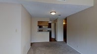This single-floor space offers a straightforward and functional layout designed for comfortable everyday living. The main living area is finished with wall-to-wall carpeting in a neutral tone, creating a warm and understated atmosphere. A textured ceiling with flush-mounted lighting provides a clean overhead finish throughout the space.

The kitchen is positioned adjacent to the living area and features a pass-through counter that connects the two spaces, promoting an open feel. Dark wood cabinetry lines the upper and lower sections of the kitchen, contrasting with light-colored countertops. A white refrigerator and standard under-cabinet appliances complete the kitchen setup, while a double sink sits beneath a window, allowing for natural light.

A hallway visible from the living area leads to additional rooms, suggesting a well-organized floor plan that separates the living and private spaces. The walls throughout are finished in a soft, neutral beige, contributing to a calm and cohesive interior palette. The overall design is practical and unassuming, with a focus on efficient use of space. The unit is well-suited for those seeking a no-frills, move-in-ready environment with a clean and functional aesthetic.