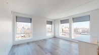 This single-floor unit showcases a clean, modern design characterized by crisp white walls, light wood-tone flooring, and an abundance of natural light throughout. The open-plan living and kitchen area is a standout feature, with oversized windows fitted with roller shades that frame expansive urban views of the surrounding cityscape. The kitchen is well-appointed with white flat-front cabinetry extending to the ceiling, a white tile backsplash, stainless steel appliances including a refrigerator, electric range, and dishwasher, and a central island with a light countertop providing additional workspace and storage. Track lighting illuminates the kitchen area effectively. The living space flows seamlessly from the kitchen, benefiting from corner window placement that maximizes light and views. The bedrooms continue the clean aesthetic, featuring neutral carpet or hardwood flooring and city-facing windows. Closet space is provided with built-in shelving and hanging storage. The bathrooms maintain the unit's contemporary palette with light tile flooring, white cabinetry, and a glass-enclosed walk-in shower finished with white subway tile in the primary bath, alongside a second full bathroom with similar clean finishes throughout.