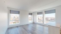 This single-floor unit showcases a clean, modern design characterized by crisp white walls, light wood-tone flooring, and an abundance of natural light throughout. The open-plan living and kitchen area is a standout feature, with oversized windows fitted with roller shades that frame expansive urban views of the surrounding cityscape. The kitchen is well-appointed with white flat-front cabinetry extending to the ceiling, a white tile backsplash, stainless steel appliances including a refrigerator, electric range, and dishwasher, and a central island with a light countertop providing additional workspace and storage. Track lighting illuminates the kitchen area effectively. The living space flows seamlessly from the kitchen, benefiting from corner window placement that maximizes light and views. The bedrooms continue the clean aesthetic, featuring neutral carpet or hardwood flooring and city-facing windows. Closet space is provided with built-in shelving and hanging storage. The bathrooms maintain the unit's contemporary palette with light tile flooring, white cabinetry, and a glass-enclosed walk-in shower finished with white subway tile in the primary bath, alongside a second full bathroom with similar clean finishes throughout.