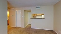 This single-floor unit features a practical and functional layout designed to maximize usable living space. The main living area is finished with wall-to-wall carpeting in a neutral tone, creating a warm and understated foundation throughout. Beige walls and white trim contribute to a clean, cohesive aesthetic that lends itself well to a variety of furnishing styles.

A defining feature of this space is the kitchen pass-through, which opens directly into the living area via a raised bar counter finished with a dark laminate surface. This design element creates a semi-open connection between the two spaces while maintaining a degree of separation. The kitchen is equipped with warm oak-toned cabinetry, offering ample storage, and a glimpse of white appliances is visible from the living area.

A doorway from the living room leads into what appears to be a hallway providing access to additional rooms. A closet with a white panel door is conveniently located near the living area, offering practical storage. The textured popcorn ceiling and centrally positioned HVAC vent are consistent throughout, and the overall layout reflects a straightforward, efficient use of space.