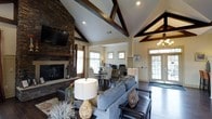 Located at 300 Cosgrove Avenue in Chapel Hill, NC, this single-story residence makes a striking first impression with its dramatic vaulted ceilings adorned with bold, dark-stained exposed wood beams that draw the eye upward and lend a sense of grandeur to the interior. The open-concept layout seamlessly connects the main living areas, creating a fluid and functional flow throughout the home. The living room serves as the centerpiece, anchored by a floor-to-ceiling stacked stone fireplace that adds both warmth and rustic character. Rich hardwood flooring runs throughout, complementing the neutral, warm-toned walls and contributing to the home's inviting ambiance. A dedicated dining area sits adjacent to the living space, offering an ideal setting for entertaining, while a built-in workspace nearby adds practical versatility. Natural light pours in through multiple windows, brightening the interior and enhancing the open feel. The entryway features elegant glass-panel doors that welcome guests with a sense of arrival. A chandelier adds a refined touch, balancing the home's blend of rustic and classic design elements. Overall, this home delivers a cohesive, well-appointed living experience with architectural details that set it apart.