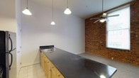 The Corner Lofts showcases a compelling blend of industrial character and contemporary design. The open-concept layout makes excellent use of its single floor, creating a spacious and airy feel throughout. A defining architectural feature is the striking exposed brick wall, which adds warmth, texture, and a nod to the building's historic character, contrasting beautifully with the clean white walls and modern finishes.

The kitchen area is well-appointed with a generous dark granite-style countertop that extends as a long bar surface, offering ample workspace and casual dining potential. Light maple cabinetry runs beneath the counter, providing substantial storage. A stainless steel sink and tile flooring complete the functional kitchen space.

Overhead, pendant lighting hangs elegantly above the kitchen counter, casting focused illumination over the workspace. A ceiling fan in the adjoining area enhances air circulation while adding to the loft's relaxed urban ambiance. The soaring ceiling height amplifies the sense of openness and volume, a hallmark of classic loft-style living.

Overall, The Corner Lofts delivers an inviting industrial-chic atmosphere, combining raw architectural details like exposed brick with polished modern fixtures to create a distinctive and functional living space.