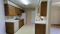This single-story space centers around a galley-style kitchen that runs along two parallel walls, creating an efficient and functional cooking area. The kitchen is lined with warm-toned wooden cabinetry on both the upper and lower sections, providing generous storage throughout. White appliances include a coil-top electric range with a ventilation hood above, a full-size refrigerator, and a dishwasher positioned beneath the countertop beside the sink. The countertops are white, offering a clean contrast against the wooden cabinetry. A double-basin sink with a black faucet is positioned along the left wall, accompanied by additional base and upper cabinets. The flooring throughout the kitchen is a light-toned vinyl tile, which is practical and easy to maintain. Overhead, a fluorescent light fixture provides bright, even illumination across the space. The textured ceiling adds a classic architectural touch. At the far end of the kitchen, a doorway leads to an adjacent room, suggesting a straightforward and connected single-floor layout. Overall, the space presents a no-frills, utilitarian design with a focus on functionality and storage capacity.
