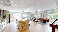 Located at 7689 Hardwood Ave S in Cottage Grove, MN, this single-floor community space offers a generously sized, open-concept layout designed for both relaxation and social gatherings. The interior is anchored by a stunning kitchen featuring a large waterfall-edge marble island with bar seating, white shaker cabinetry with brass hardware, a glittering mosaic tile backsplash, stone countertops, stainless steel appliances including a French door refrigerator, and a wall-mounted television — making it as functional as it is visually impressive. Light wood-look flooring flows throughout, creating a warm, cohesive aesthetic that ties the entire space together. The lounge area is thoughtfully appointed with a rich leather sofa, mid-century modern accent chairs in warm tones, a shuffleboard table, and a large wall-mounted display screen for entertainment. Tray ceilings with recessed lighting and an elegant circular chandelier add architectural character overhead. Abundant natural light pours in through oversized windows on multiple walls, offering lush treetop views, while large sliding glass doors connect the interior directly to an outdoor terrace furnished for al fresco dining and entertaining. The overall ambiance is modern, inviting, and well-curated.