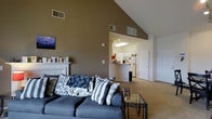This single-floor residence showcases an open and airy layout defined by its dramatic vaulted ceilings, which lend a generous sense of height and volume to the main living area. The living room serves as the heart of the home, featuring a classic white fireplace with a mantle, warm neutral-toned walls, and soft carpeted flooring that adds comfort underfoot. The space is well-ventilated with ceiling vents positioned throughout, ensuring consistent climate control.

The living area flows seamlessly into the kitchen, which is visible through an open pass-through layout. The kitchen features white cabinetry, a refrigerator, and a functional peninsula counter that separates it from the main living space while maintaining an open connection — ideal for entertaining or casual interaction. A skylight above the kitchen enhances the brightness of the space with natural overhead light.

Multiple interior doors lead to additional areas of the home, suggesting private spaces such as bedrooms or utility areas that branch off from the central hub. The overall color palette is understated and cohesive, with taupe and white tones creating a calm and neutral ambiance throughout. The combination of high ceilings, open-plan design, and abundant light makes this a welcoming and functional single-floor home.