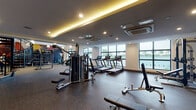 This expansive fitness center is thoughtfully designed to accommodate a full range of workout needs across a single, open floor plan. The space is divided into distinct functional zones, each outfitted with professional-grade equipment. A row of treadmills and StairMaster machines lines the perimeter near large floor-to-ceiling windows, offering natural light and scenic outdoor views of surrounding greenery and water. A dedicated strength training area features a comprehensive dumbbell rack, adjustable benches, and cable machines, while a separate TRX Training Zone is equipped with battle ropes, suspension trainers, and additional functional fitness tools.

Architecturally, the facility stands out with soaring double-height ceilings in the training zone section, pendant lighting, and a striking spiral staircase serving as a design focal point. A vibrant geometric mural in bold reds, yellows, and charcoals adds visual energy to the space. The opposite section features a dropped ceiling with warm amber cove lighting and recessed spotlights, creating a more intimate ambiance. Durable rubberized flooring runs throughout, and built-in shelving and a workstation near the staircase add functional convenience. Climate control is provided via ceiling-mounted air conditioning units.