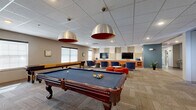 Located at 1521 Sixth Ave in Troy, NY, this single-floor amenity center offers a generously sized, thoughtfully designed space built to serve a variety of community needs. The open-plan main lounge is an expansive gathering area anchored by comfortable sectional seating, a pool table, and multiple seating clusters, including high-top bar tables near the entrance and intimate round tables throughout. A decorative wall mural featuring a river motif adds an artistic, locally inspired touch to the lounge's neutral color palette of warm tans, oranges, and teal accents. A vending area with snack and beverage machines provides convenient refreshment access alongside vibrant orange and white casual seating. A dedicated TV lounge features bold orange cabinetry, a mounted flat-screen television, and plush blue armchairs, creating a cozy media retreat. The facility includes a well-equipped fitness center with cardio machines, free weights, cable equipment, floor mats, and full-wall mirrors, complemented by rubberized flooring in a colorful striped design. A classroom-style meeting room accommodates group sessions with long folding tables, chairs, and wall-mounted whiteboards, while a smaller conference room offers a round table, projector, whiteboard, and individual climate control. Two bathrooms complete the layout, ensuring full functionality throughout this versatile community amenity space.