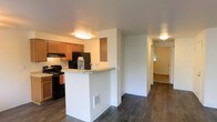 This single-story space showcases an open and functional layout with dark hardwood-style flooring that extends throughout the main living areas, creating a cohesive and clean aesthetic. The kitchen is equipped with warm honey-toned wood cabinetry, black appliances including a range and refrigerator, and an under-cabinet range hood. A white peninsula with a light-colored countertop serves as a breakfast bar, providing both additional prep space and a casual dining option. Flush-mount ceiling light fixtures provide consistent ambient lighting across the space. A half-wall divides the kitchen from the adjacent hallway, which leads to additional rooms visible in the background. Natural light filters in from a window at the far end of the hallway, brightening the deeper areas of the unit. The neutral white walls complement the darker flooring and warm cabinetry tones, resulting in a balanced and inviting interior. The open sightlines between the kitchen and the rest of the living space contribute to an airy feel, making the overall floor plan feel connected and easy to navigate.