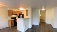 This single-story space showcases an open and functional layout with dark hardwood-style flooring that extends throughout the main living areas, creating a cohesive and clean aesthetic. The kitchen is equipped with warm honey-toned wood cabinetry, black appliances including a range and refrigerator, and an under-cabinet range hood. A white peninsula with a light-colored countertop serves as a breakfast bar, providing both additional prep space and a casual dining option. Flush-mount ceiling light fixtures provide consistent ambient lighting across the space. A half-wall divides the kitchen from the adjacent hallway, which leads to additional rooms visible in the background. Natural light filters in from a window at the far end of the hallway, brightening the deeper areas of the unit. The neutral white walls complement the darker flooring and warm cabinetry tones, resulting in a balanced and inviting interior. The open sightlines between the kitchen and the rest of the living space contribute to an airy feel, making the overall floor plan feel connected and easy to navigate.