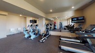 This fitness center is a thoughtfully designed, single-floor facility built to support a comprehensive workout experience. The space is accessed through a dedicated entrance area featuring neutral-toned walls, recessed lighting, carpeted flooring, and a small seating arrangement — offering a clean, welcoming transition into the gym.

The main workout floor is generously proportioned and finished with durable rubber flooring suited for high-impact activity. A full-length wall mirror spans one side of the room, visually expanding the space and providing useful feedback during exercise. Recessed ceiling lights provide consistent, even illumination throughout.

The cardio zone is well-stocked with multiple treadmills, elliptical trainers, a stationary bike, and a recumbent bike, positioned near large windows fitted with horizontal blinds that admit natural light and offer views to the exterior. Mounted flat-screen televisions are available for entertainment during workouts.

Strength and functional training equipment includes a multi-station cable machine with a bench, a rack of stability balls in varying sizes, and open floor space suitable for stretching or bodyweight exercises. Framed artwork adorns the walls, adding a polished, community-oriented aesthetic to the environment. The overall ambiance is clean, functional, and well-maintained, making it a practical and inviting space for daily fitness routines.
