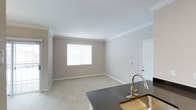This single-floor space presents a clean, open-concept layout that seamlessly connects the kitchen and living areas. The living room is finished with plush wall-to-wall carpeting in a light neutral tone, contributing to a warm and comfortable atmosphere. Soft gray walls are complemented by crisp white crown molding that frames the ceiling, adding a subtle architectural detail that elevates the overall aesthetic. A window fitted with white horizontal blinds allows natural light to filter into the living area, brightening the space while maintaining privacy.

The kitchen, visible in the foreground, features a dark countertop surface that provides a striking contrast against the lighter tones of the surrounding space. A brushed gold pull-down faucet serves as a stylish focal point above a gold-toned sink, reflecting a cohesive modern design sensibility. A white paneled door provides access to the exterior, adding functionality to the layout. The consistent use of neutral colors and clean lines throughout creates a cohesive, move-in-ready environment that is both practical and visually balanced.