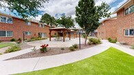 Located at 2112 Moon St NE in Albuquerque, New Mexico, this residential complex is defined by its classic two-story brick construction with a warm, earthy tone complemented by blue steel railings and stairways. The buildings are arranged around a series of well-maintained communal courtyards that serve as the heart of the property. Mature trees are interspersed throughout the grounds, providing shade and a sense of established character to the outdoor spaces.

The courtyards feature a mix of lush green turf areas, gravel landscaping with drought-tolerant native plantings, and wide concrete walkways that connect the various building entrances. A standout feature of the shared outdoor space is a wood-framed gazebo with picnic seating, creating an inviting gathering area for residents. Additional seating, including exterior benches, is positioned throughout the grounds.

Each building level is accessible via sturdy blue steel staircases, and upper-level units are appointed with private balconies enclosed by matching blue railings. Ground-floor units open directly onto landscaped walkways, with large windows allowing natural light into the interiors. The overall ambiance of the property is functional and community-oriented, with thoughtfully maintained grounds that reflect a blend of Southwestern landscaping sensibility and practical residential design.
