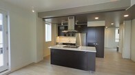 This single-floor space showcases a contemporary, open-concept layout centered around a well-appointed kitchen. The kitchen is a standout feature, designed with rich dark cabinetry that contrasts sharply against crisp white countertops on the central island, which serves as both a workspace and a visual anchor for the room. A stainless steel island range hood rises prominently above the cooking area, adding an industrial yet refined touch to the space. Full-height dark cabinetry lines the back wall, providing ample storage, while stainless steel appliances, including a refrigerator, integrate seamlessly into the design. The kitchen island includes a built-in cooktop, enhancing the functionality of the space. Light-toned hardwood flooring runs throughout, offering a warm counterbalance to the darker cabinetry and lending the space an airy feel. Recessed lighting and track lighting illuminate the kitchen effectively, while a window allows natural light to filter into the adjoining area. The open layout connects the kitchen to the living area, creating a cohesive and fluid living environment with a clean, modern aesthetic throughout.