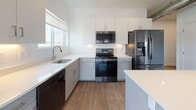 This contemporary single-floor unit showcases a clean, modern aesthetic throughout, defined by crisp white walls, recessed lighting, and a cohesive design language. The open-concept living and dining area flows seamlessly into the kitchen, where rich hardwood-style flooring adds warmth to the bright space. Large windows and a sliding glass door to a private balcony flood the living area with natural light and offer views of the surrounding greenery.

The kitchen is well-appointed with white upper cabinetry, light grey lower cabinets, white quartz countertops, a center island with pendant lighting, and a full suite of stainless steel appliances including a refrigerator, electric range, microwave, and dishwasher. A window above the sink further enhances the bright, airy feel.

A notable architectural detail is the exposed metallic ductwork that runs through the open living area and hallway, lending an industrial-chic character to the otherwise minimal interior.

The bedrooms feature plush carpet flooring and generous natural light. One bedroom includes a walk-in closet with direct views into the kitchen area. Both bathrooms are finished with grey cabinetry, gold-toned hardware, quartz countertops, and subway tile — one with a tub/shower combo and one with a walk-in shower. A dedicated in-unit laundry closet with a stacked washer and dryer completes the space.