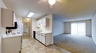This single-floor residence offers a practical and functional layout designed for comfortable everyday living. The kitchen is outfitted with white cabinetry, a double-basin sink, a dishwasher, and a refrigerator, illuminated by a skylight that brings in natural overhead light. Tile flooring runs throughout the kitchen area, providing a clean and durable surface. The kitchen flows seamlessly into an open living area, which features plush carpet flooring and a ceiling fan overhead. A sliding glass door at the far end of the living space allows natural light to filter in and provides direct access to the outdoors. A hallway visible from the kitchen leads to additional rooms, including what appears to be a bedroom with mirrored closet doors and a bathroom, offering a well-connected and easy-to-navigate floor plan. The overall ambiance is neutral and understated, with light-toned walls throughout that create a clean, open atmosphere suited to a variety of interior styles.