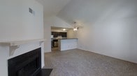 This single-floor residence offers a thoughtfully designed open-concept layout that seamlessly connects the living and kitchen areas. The living room is highlighted by soaring vaulted ceilings that lend an expansive, airy quality to the space, while plush carpeting underfoot adds warmth and comfort. A classic fireplace with a clean white mantel serves as a natural focal point, making the area inviting and well-anchored. A ceiling fan is centrally positioned to ensure year-round comfort throughout the space. The kitchen is accessed through an open passthrough and features rich, dark cabinetry that contrasts crisply against light walls, a dark countertop peninsula ideal for casual dining or prep work, and stainless steel appliances that add a modern, functional touch. The peninsula also provides a subtle visual boundary between the kitchen and living areas while preserving the open flow of the layout. Neutral tones throughout — from the soft carpet to the white walls — create a clean, versatile backdrop ready for personalization. The overall ambiance is bright, open, and welcoming, with architectural details like the vaulted ceiling adding character and a sense of generous scale.
