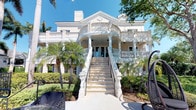 This distinguished estate showcases an impressive neoclassical architectural design that commands immediate attention. The grand façade is finished in crisp white, featuring an elegant curved portico supported by stately columns that frame the main entrance. A sweeping dual staircase with decorative balusters leads up to the covered entryway, lending a sense of ceremony and grandeur to arrivals. Above, a wraparound second-floor balcony with matching ornamental railings extends across the front of the home, offering elevated views of the surrounding grounds. Detailed cornice work, pediment accents, and classical moldings are showcased throughout the exterior, reflecting a meticulous attention to architectural craftsmanship. The property is enveloped by lush tropical landscaping, with mature palm trees, manicured hedges, and vibrant greenery that complement the bright white structure beautifully. The outdoor areas include generous patio space suitable for entertaining, with glimpses of stylish outdoor furnishings. Natural light floods the exterior through multiple sets of glass French doors, hinting at bright, airy interiors within. The overall ambiance is one of refined elegance, tropical luxury, and timeless architectural beauty.