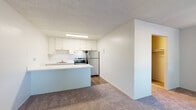 This single-floor unit offers a practical and functional layout designed for comfortable everyday living. The kitchen is well-appointed with white cabinetry featuring modern hardware, light countertops, a stainless steel sink, an electric range, and a full-size refrigerator. A peninsula counter extends into the adjacent living area, creating a natural division between the two spaces while maintaining an open feel. The living area is finished with plush carpeting and features a large window dressed with vertical blinds, allowing natural light to fill the room. A wall-mounted heating unit provides efficient climate control. Transitioning toward the sleeping area, a generous walk-in closet offers ample storage with a built-in hanging rod. The bathroom is clean and bright, finished with white tile surrounds and equipped with a full soaking tub and a multi-function handheld showerhead with chrome fixtures. Throughout the unit, textured ceilings and neutral wall tones create a cohesive, understated aesthetic. The combination of hard-surface flooring in the kitchen and soft carpet in the living and sleeping areas adds both practicality and comfort to the overall design of the space.