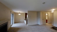 This single-floor space offers an open-concept layout that seamlessly connects the living area to the kitchen. The interior is finished with neutral beige walls and wall-to-wall carpeting, creating a clean and cohesive aesthetic throughout. The living area is generously sized, receiving natural light from a large sliding glass door along one wall. A pendant light fixture serves as a subtle focal point, adding warmth to the space. A floor vent indicates a built-in heating system for year-round comfort. The kitchen is partially separated by a half-wall counter with a breakfast bar overhang, providing both a functional workspace and an informal dining or seating area. Dark cabinetry in the kitchen contrasts with the lighter tones of the surrounding walls, and a window above the sink allows natural light into the cooking area. A thermostat is mounted on the wall, centrally located for easy access. A paneled door on the far side of the space leads to an additional room. The overall ambiance is clean, neutral, and move-in ready, with a practical and straightforward floor plan.