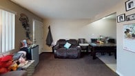 This single-story residence offers a functional layout across several rooms, each finished with textured ceilings and neutral-toned walls. The living room is a comfortable gathering space featuring dark brown carpet, a bold chocolate-brown accent wall, and a large flat-screen television on a media console. Two sizable dark upholstered sofas provide ample seating, and framed wall art adds a personal touch to the space.

The kitchen is equipped with rich dark wood cabinetry extending to the upper walls, granite-style countertops forming an L-shaped workspace, and black appliances including an electric range and built-in microwave hood. A secondary living or dining area adjoins the kitchen, furnished with a reclining loveseat and a dark dining table set.

Two bedrooms are present. The primary bedroom features a ceiling fan with light fixture, a large mirrored sliding-door closet, a window offering natural light and outdoor views, and carpeted floors. A second bedroom accommodates a large flat-screen TV, a painted dresser, and a baby crib. The bathroom includes a dark wood vanity with a granite-style countertop, an oval mirror with overhead lighting, and tile flooring.