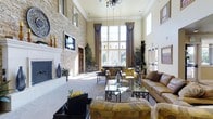 This elegant single-floor clubhouse offers a grand and welcoming atmosphere defined by its dramatic double-height ceilings and expansive open floor plan. The main gathering area is anchored by a striking stacked stone fireplace wall, which adds warmth and texture as a central architectural focal point. A wall-mounted television is integrated seamlessly into the stone surround, enhancing the space's functionality.

Floor-to-ceiling windows dressed with rich, floor-length drape panels flood the interior with abundant natural light while providing views of the surrounding neighborhood. The soaring ceiling height is further accentuated by an ornate chandelier that adds a refined, classic touch to the overall design.

The space is thoughtfully furnished with a mix of plush leather sofas, patterned accent chairs, and glass-topped coffee tables, creating comfortable seating arrangements suited for both casual socializing and organized events. A billiards table is visible toward the rear of the space, adding a recreational element to the clubhouse experience.

Lush indoor plants are strategically positioned throughout, lending a fresh, vibrant quality to the interior. The neutral carpet, cream-toned walls, and warm wood accents work together to create an inviting, cohesive aesthetic that is both sophisticated and approachable — ideal for a community gathering destination.