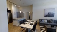 This single-story space in Carrollton, Texas, showcases a thoughtfully designed open-concept layout that connects the kitchen, dining, and living areas into one cohesive and functional environment. The soaring ceilings amplify the sense of space and airiness throughout, while light wood-tone flooring runs consistently underfoot, lending warmth and visual continuity to the interior.

The kitchen features a peninsula-style counter with granite or stone-look countertops and an undermount sink, offering both prep space and a casual seating area with bar-height chairs. Pendant lighting hangs above the peninsula, providing focused illumination with a contemporary aesthetic, complemented by track lighting overhead.

Adjacent to the kitchen, the dining area accommodates a rectangular table with seating for four to six, set beneath additional pendant fixtures. The setup is ideal for both everyday meals and entertaining. Just beyond, the living area is anchored by a gray upholstered sofa accented with decorative pillows, resting atop a bold striped area rug that adds a graphic element to the space.

Neutral gray walls and trim create a clean, modern backdrop throughout, while a hallway visible in the background suggests access to additional private rooms, completing this well-appointed and cohesive layout.