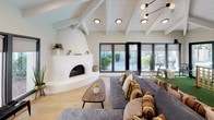 This well-appointed single-floor space blends Southwestern charm with modern design. The living area is defined by striking vaulted ceilings with exposed white-painted wood beams, a sculptural kiva-style fireplace with a smooth plaster surround, and light hardwood flooring. Walls of large, dark-framed windows and sliding glass doors flood the space with natural light and provide panoramic views of the surrounding outdoor areas, including a pool patio with seating. A modern sputnik-style pendant light adds a mid-century touch above the generously sized seating area.

Adjacent to the living space is a dedicated games zone, featuring artificial turf flooring and a jumbo Connect Four game, creating a playful and interactive environment. This area offers additional access to the outdoor spaces through its own set of sliding glass doors.

The kitchen features an expansive countertop in white quartz with bold grey veining, complemented by rich walnut cabinetry with glass-panel accents. Striped Roman shades add a warm, colorful touch, while desert-themed artwork and a decorative cactus sculpture nod to the Southwestern aesthetic. Recessed lighting throughout ensures a bright and welcoming ambiance across all areas of the space.