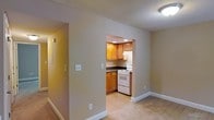 Located at 1933 North Hills Dr in Raleigh, NC, this single-floor unit offers a straightforward, functional layout with a cohesive neutral color palette throughout. The living area features light-toned carpet and smooth painted walls in a warm beige hue, creating a calm, understated ambiance. A textured ceiling adds subtle architectural character to the space.

The kitchen is accessible via an open pass-through cutout in the wall, creating a sense of visual connectivity between the living and cooking areas. Inside, the kitchen is outfitted with warm honey-toned wood cabinetry both above and below the countertops, providing ample storage. Dark laminate countertops offer a grounding contrast to the lighter cabinetry. A white electric range and vent hood are positioned along the back wall, keeping the layout compact and efficient. Recessed lighting in the kitchen provides focused illumination over the workspace.

A hallway visible from the main living area suggests additional rooms beyond, with consistent flooring and wall tones maintaining a unified feel throughout. White baseboards throughout the unit provide a clean, finished border between the walls and flooring, contributing to the overall tidy and well-maintained appearance of the space.