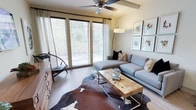This single-floor residence offers a thoughtfully designed living space that balances modern aesthetics with warm, inviting details. The living area serves as the focal point of the home, showcasing an abundance of natural light streaming through large, floor-to-adjacent-ceiling windows dressed with horizontal blinds and flowing curtains. The generous window arrangement offers pleasant views of surrounding greenery, creating a seamless connection between the interior and the outdoors. A ceiling fan with a sleek finish adds both function and visual interest overhead. The space is anchored by a contemporary sectional sofa in a neutral gray tone, complemented by an eclectic mix of throw pillows. A live-edge wood coffee table with metal legs serves as a striking centerpiece, resting atop a cowhide area rug that adds texture and character to the hardwood flooring beneath. An arc floor lamp provides warm ambient lighting, while a curated gallery of framed artwork adorns the walls, lending a refined, gallery-like quality to the room. Additional seating in the form of a sculptural accent chair rounds out the layout. Overall, the space exudes a contemporary yet comfortable ambiance, with carefully chosen design elements that create a cohesive and welcoming environment.