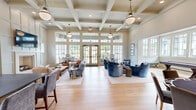 Located at 5830 Elsie Road in Birmingham, Alabama, this refined single-floor living space offers a thoughtfully designed open-concept layout that balances elegance with comfort. The room is anchored by dramatic double-height coffered ceilings adorned with globe pendant lights, creating a grand yet welcoming atmosphere throughout.

Large floor-to-ceiling windows and French doors line multiple walls, flooding the interior with natural light and offering open views of the surrounding landscape. Light-toned hardwood flooring runs throughout, reinforcing the bright and airy ambiance.

The space is organized into several distinct yet connected zones. A cozy seating area near the fireplace features a built-in tile surround, wall-mounted television, and paneled wainscoting, complemented by plush sofas and leather armchairs. A central lounge area showcases plush blue velvet swivel chairs arranged around low accent tables. A shuffleboard table and additional seating provide a recreational corner, while a dedicated dining area and a built-in bar or service counter round out the space's versatile programming.

The overall design palette is soft and neutral — warm whites, cool blues, and natural wood tones — conveying a polished, community-oriented environment ideal for gathering and relaxation.