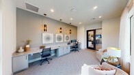 Located at 19815 Kenswick Drive in Humble, Texas, this single-floor space functions as a well-appointed business center designed to support productivity in a polished, contemporary setting. The interior is characterized by a neutral color palette of soft grays and warm whites, complemented by light-toned carpet flooring that runs throughout.

The primary workspace features an expansive built-in desk that spans the length of one wall, finished in a light surface and supported by sleek gray cabinetry below. Multiple workstations are equipped with desktop computers, providing ample space for individual use. Above the desk, a trio of framed geometric art pieces adds visual interest, while three elongated pendant lights with dark, smoked glass shades hang from the ceiling, creating a focused and sophisticated atmosphere.

Recessed lighting supplements the pendants throughout the space, ensuring consistent illumination. A separate lounge area offers comfortable upholstered seating arranged near floor-to-ceiling sheer-draped windows, which allow generous natural light and views of the surrounding outdoor landscape. Glass-paneled interior doors connect distinct areas, maintaining an open, interconnected feel while providing visual separation. The overall ambiance is professional yet inviting.