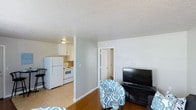 This single-floor unit offers a practical and well-organized layout designed to maximize the use of available space. The living area serves as the central hub of the unit, featuring warm wood-style flooring that adds a sense of comfort and continuity. Light gray walls paired with white baseboards create a clean, neutral backdrop, while the textured ceiling adds subtle depth to the interior. The space accommodates a sitting area with room for a television setup, making it a functional everyday living environment. Adjacent to the living area, the kitchen is efficiently arranged and outfitted with white cabinetry, a white refrigerator, and a white range, all contributing to a cohesive, bright aesthetic. Tile flooring in the kitchen provides a practical transition from the main living space. A hallway visible from the main room leads to additional areas of the unit, suggesting a thoughtful separation between the living and private spaces. Overall, the unit presents a straightforward, functional design with consistent finishes throughout, making it a comfortable and easy-to-navigate single-floor living space.